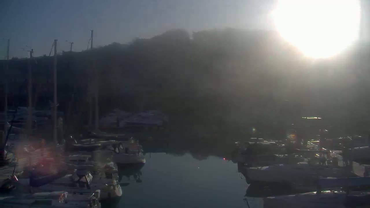 A hazy sunrise illuminates a marina filled with numerous boats moored along a calm waterway, with a hill shrouded in mist in the background.