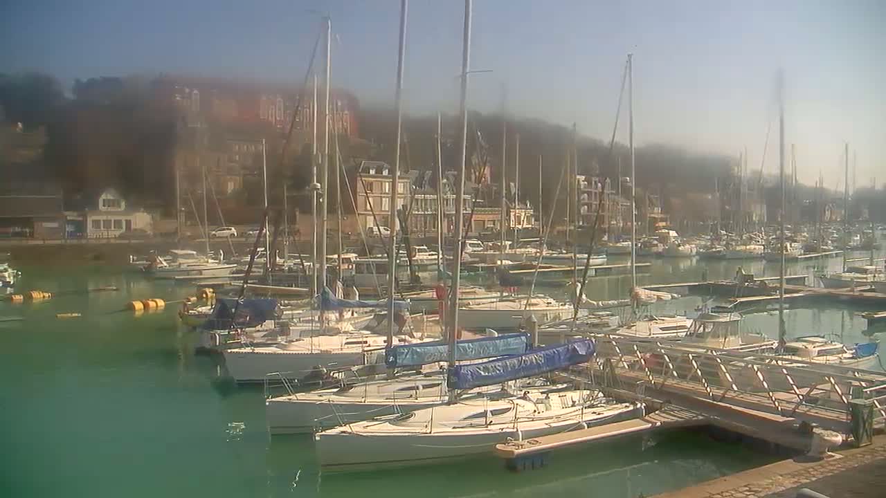 A marina filled with sailboats and motorboats on a sunny day with a hazy atmosphere over the buildings on the hillside in the background.