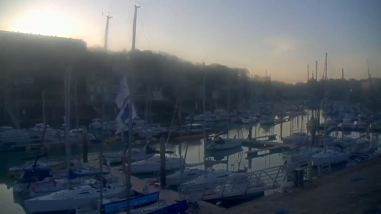 A marina filled with numerous sailboats and motorboats is shrouded in a hazy, misty atmosphere at sunrise.