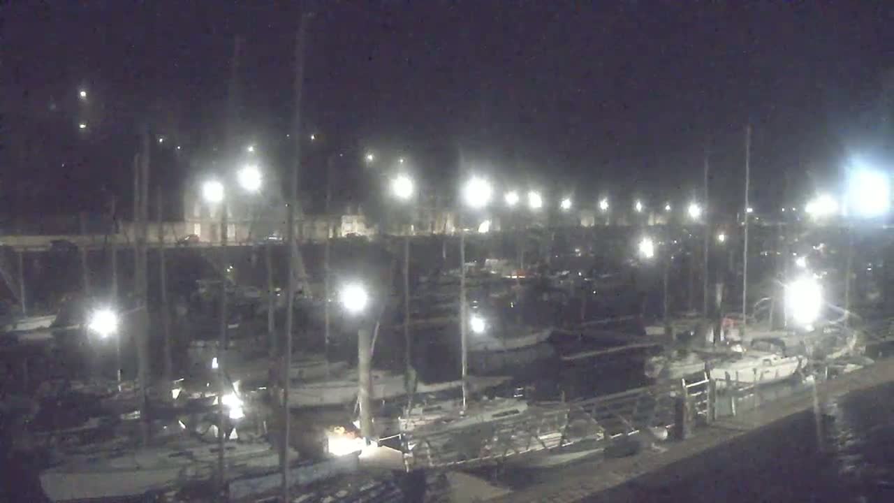 Many boats are docked at a marina at night under bright lights.