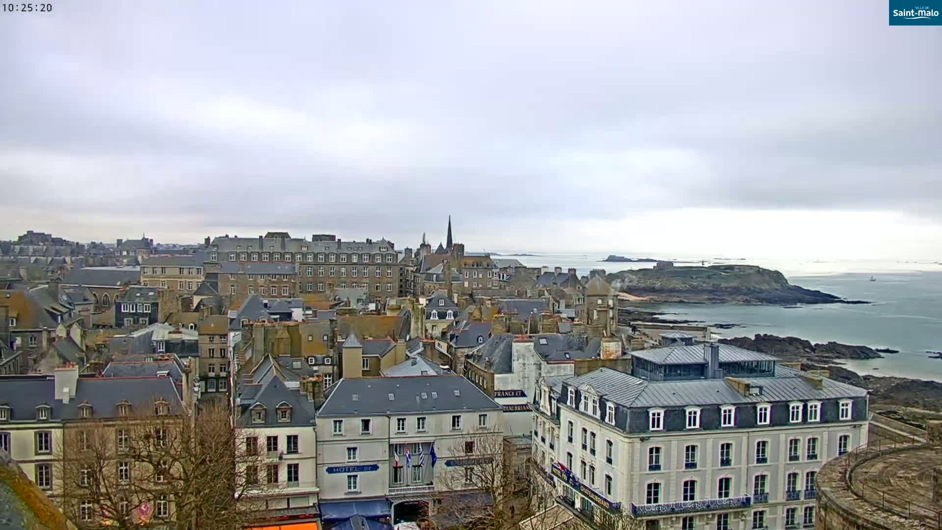 An overcast day reveals a coastal town with many grey-roofed buildings, a rocky shoreline, and a calm sea extending to the horizon.
