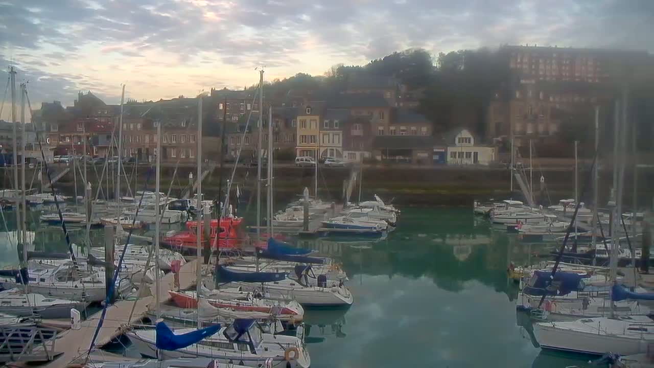 A marina filled with sailboats and motorboats is nestled next to a town under a cloudy, sunrise sky.