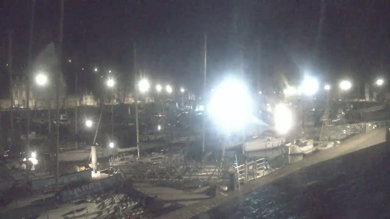 A nighttime view of a marina filled with sailboats and other vessels under bright artificial lights.