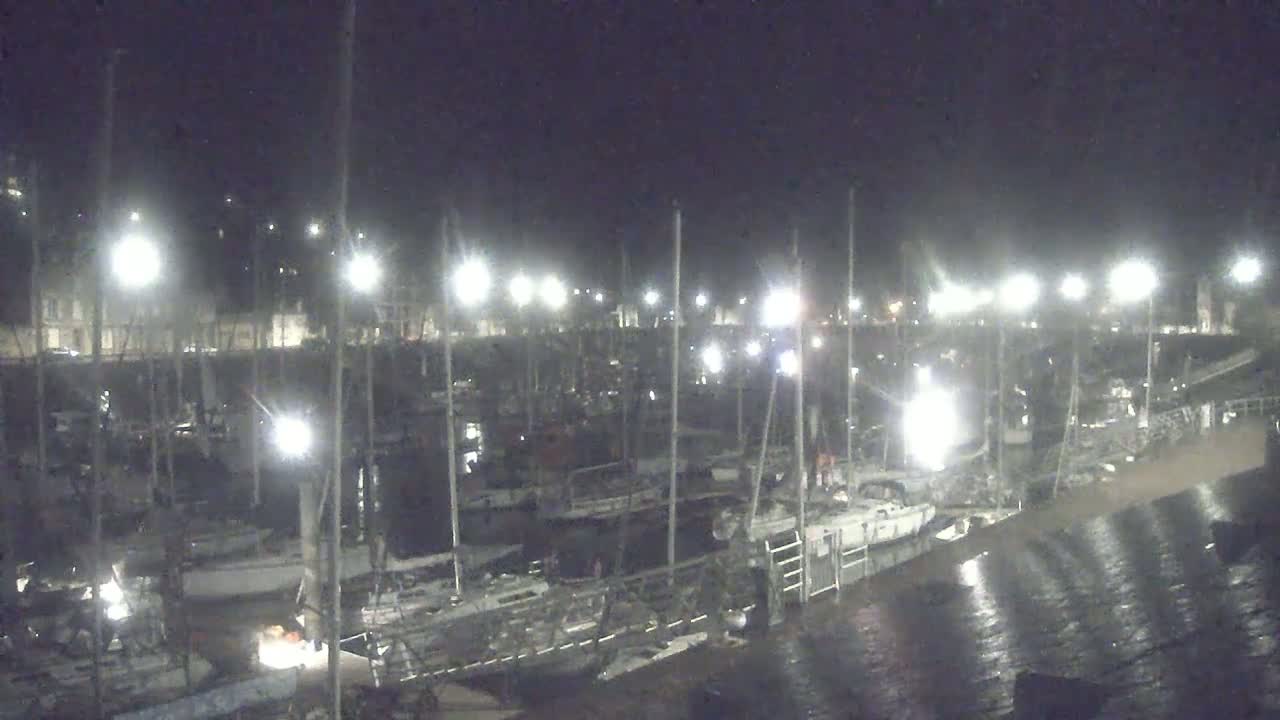 Many sailboats are docked at a marina at night under bright lights.