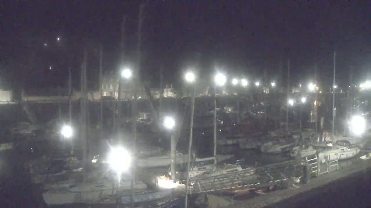 A nighttime view of a marina filled with numerous sailboats docked under bright artificial lights.