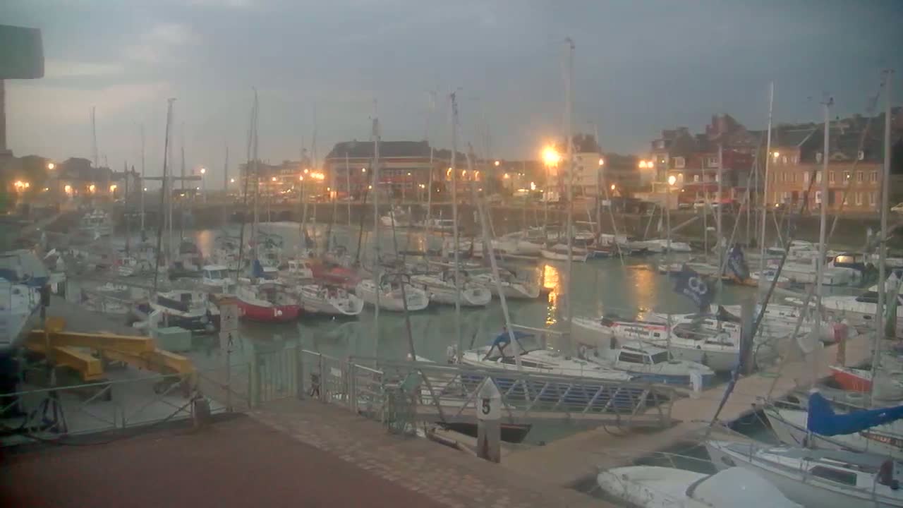Many sailboats are docked in a harbor at twilight under an overcast sky.