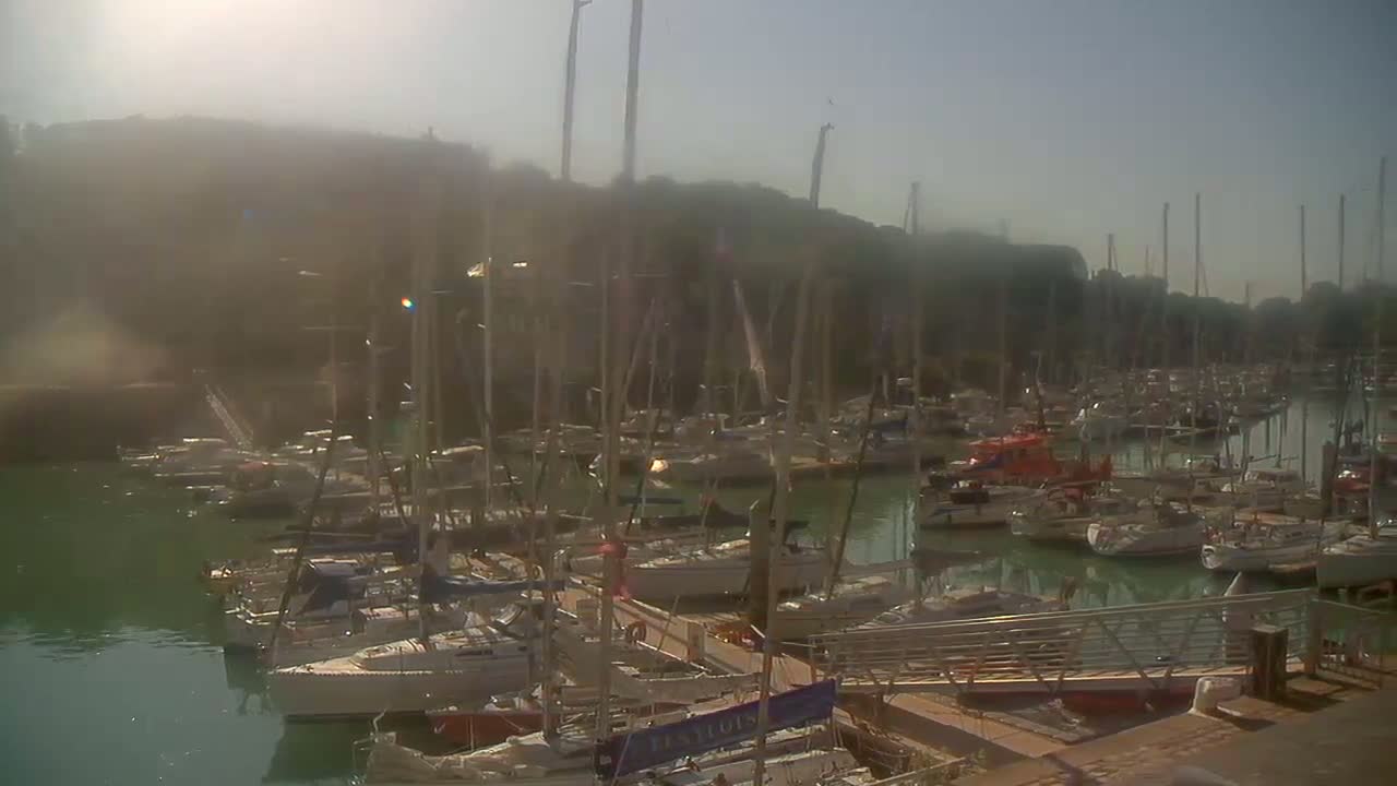 A hazy sunny day reveals a marina filled with numerous sailboats and motorboats docked along piers.