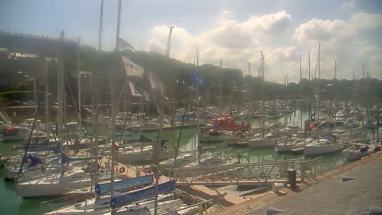 A marina filled with numerous sailboats and motorboats is seen on a partly cloudy, sunny day.