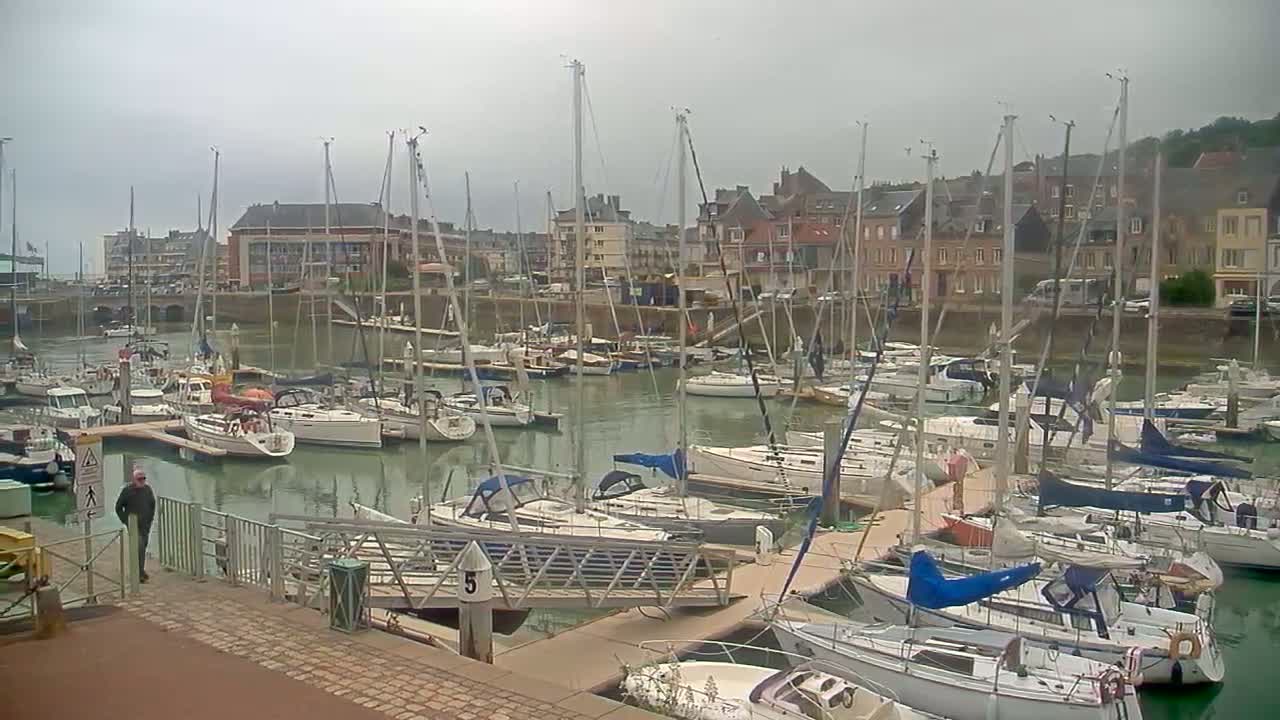 A man walks along a dock next to a marina filled with sailboats on an overcast day.