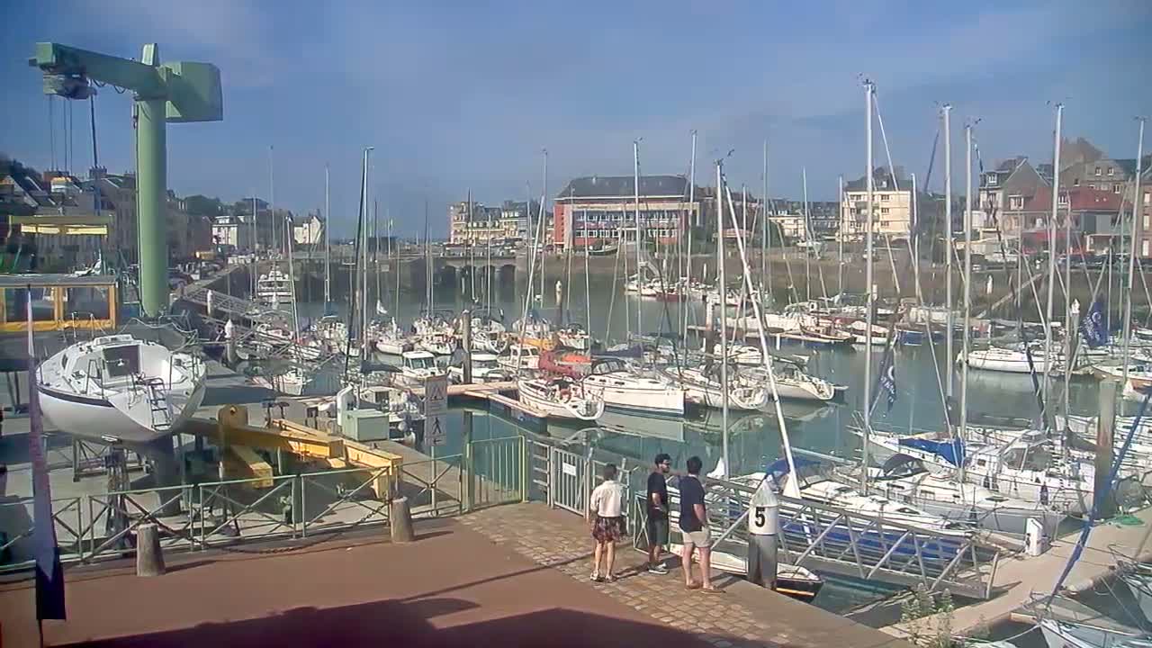 A marina filled with numerous sailboats and motorboats on a sunny day, with a few people standing near a boat lift on the dock.