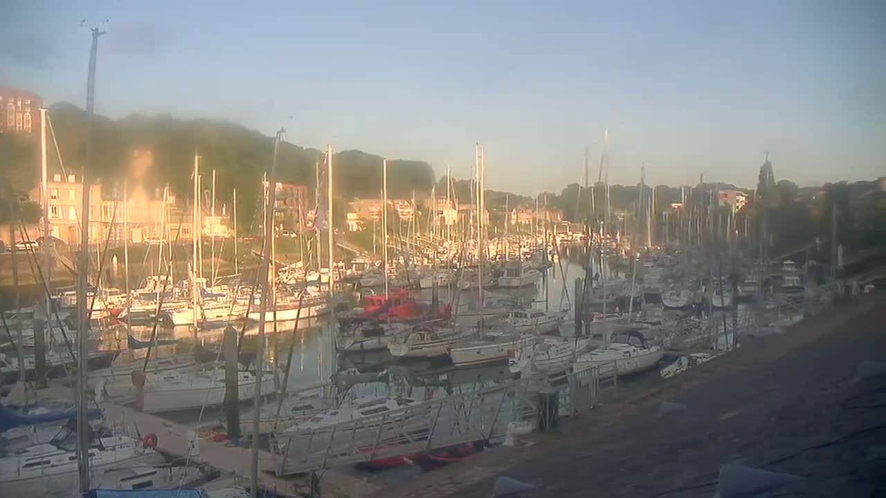 A marina filled with numerous sailboats and motorboats is seen at sunrise under a hazy sky.