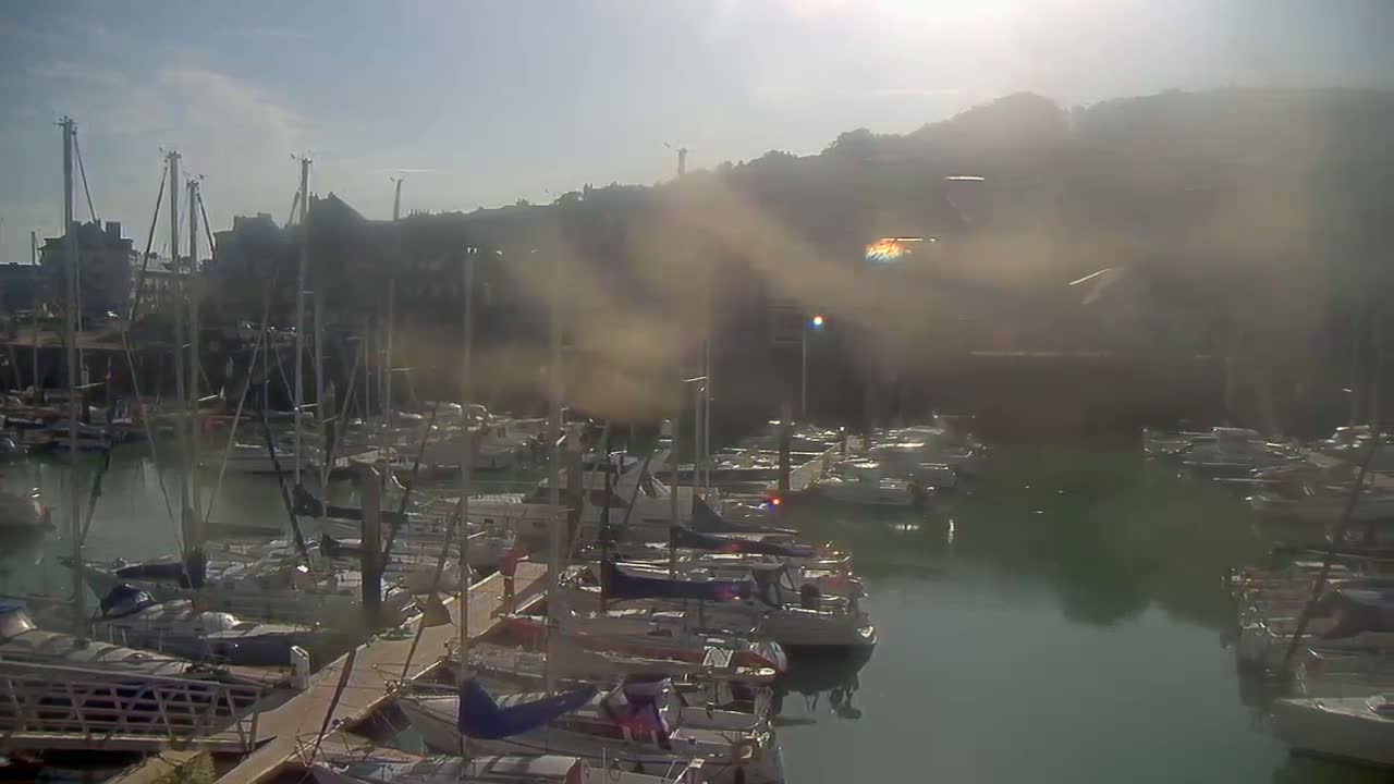 A hazy, sunny day shows a marina filled with numerous sailboats and motorboats docked along piers.