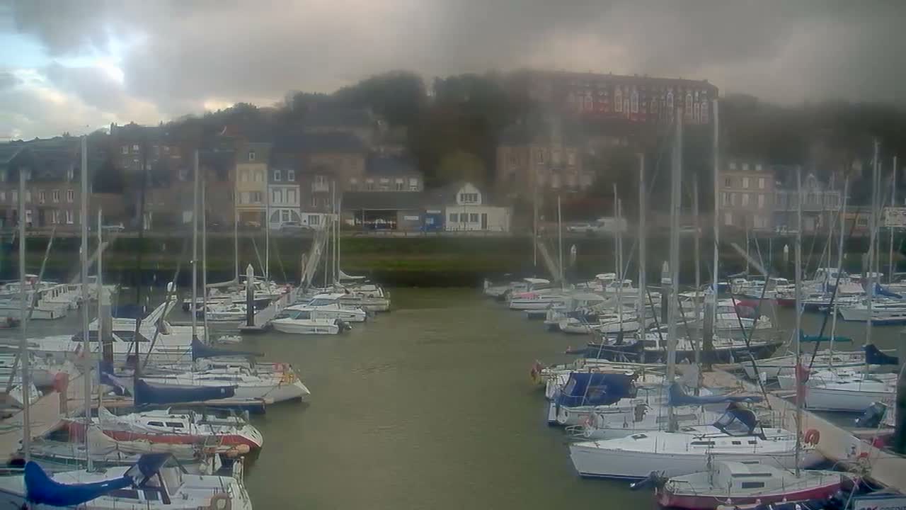 Many sailboats and powerboats are docked in a river or canal, with buildings lining the hilly banks under a heavily overcast and gloomy sky.