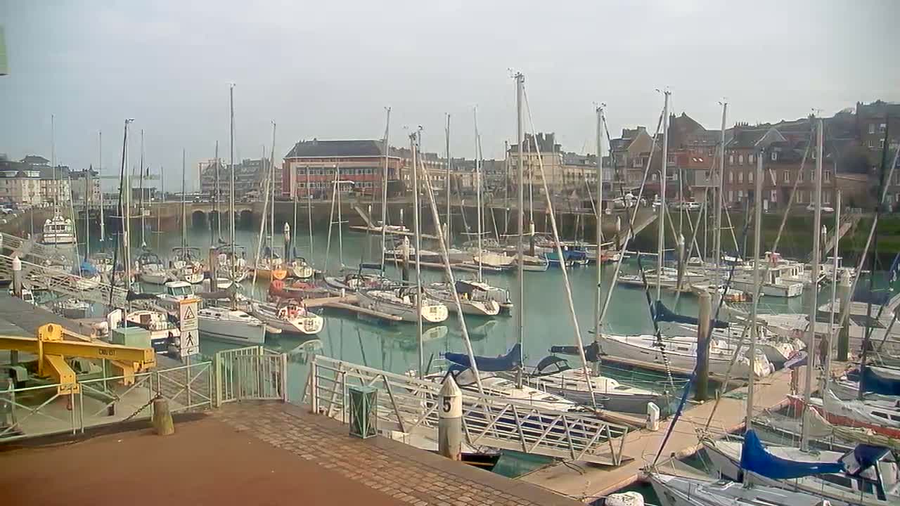 Many sailboats and powerboats are docked in a river or canal, with buildings lining the hilly banks under a heavily overcast and gloomy sky.