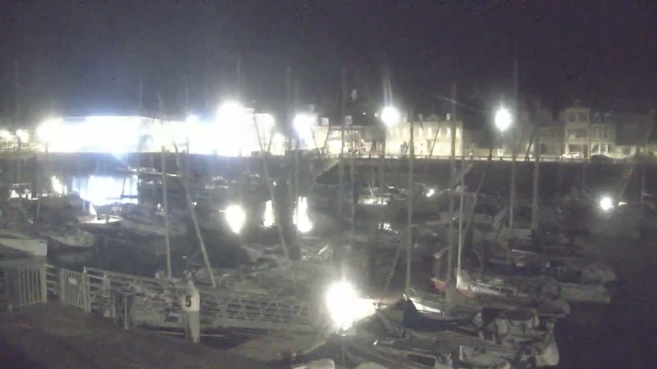 Many sailboats and powerboats are docked in a river or canal, with buildings lining the hilly banks under a heavily overcast and gloomy sky.