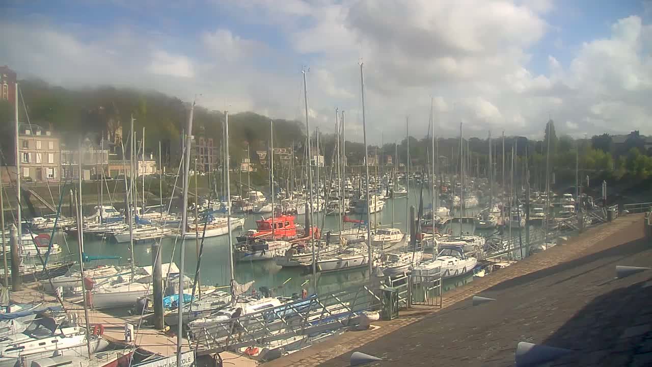 Many sailboats and powerboats are docked in a river or canal, with buildings lining the hilly banks under a heavily overcast and gloomy sky.