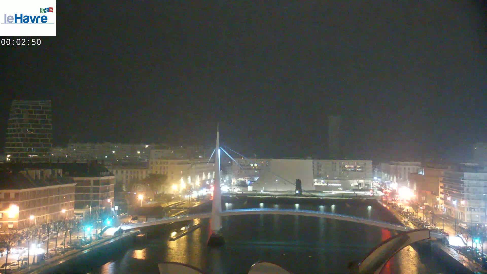 A nighttime view of a city with a modern, illuminated bridge spanning a waterway, under an overcast sky.