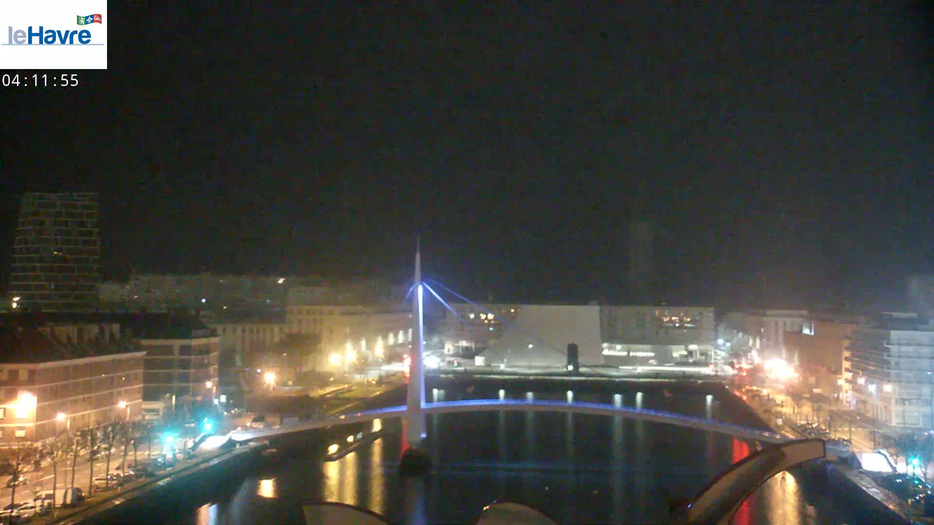 A nighttime view of a city with a modern bridge illuminated in blue over a calm waterway, under a clear, dark sky.