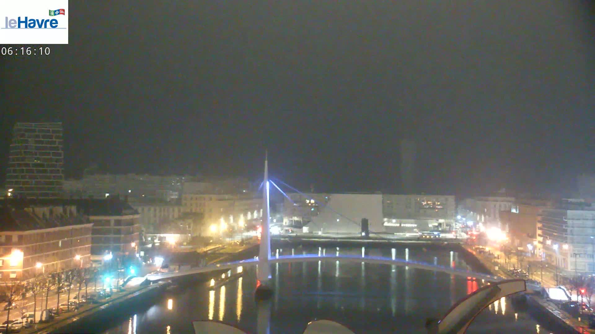 A nighttime view of a city harbor with a lit bridge spanning a calm body of water under hazy conditions.