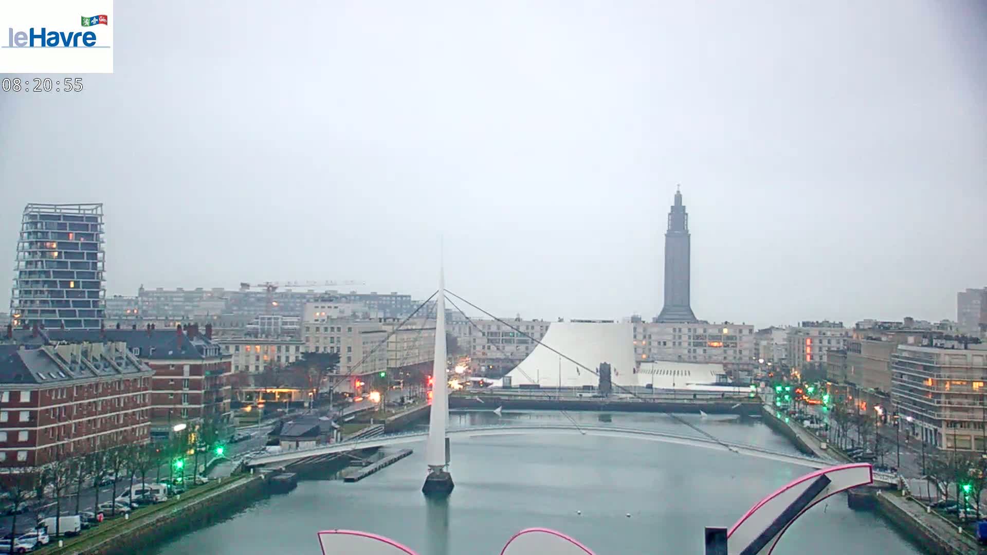 A city landscape on an overcast day shows a waterway with a slender bridge, buildings lining the water's edge, and a tall tower in the distance.