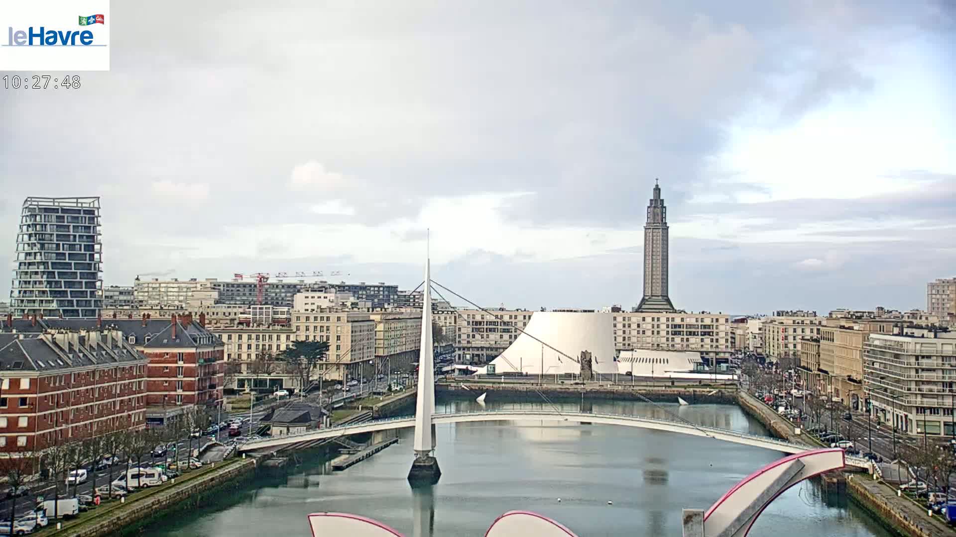 A cityscape with a modern white bridge over a calm waterway, under a mostly cloudy sky, features several buildings including a tall tower and a domed structure.