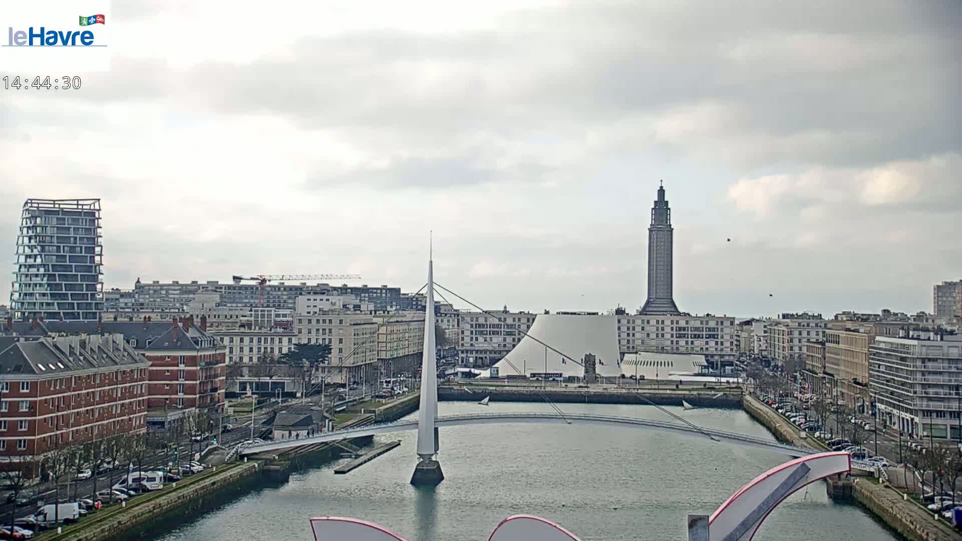 A cityscape with overcast skies shows a body of water, a modern bridge, and various buildings, including a tall tower.