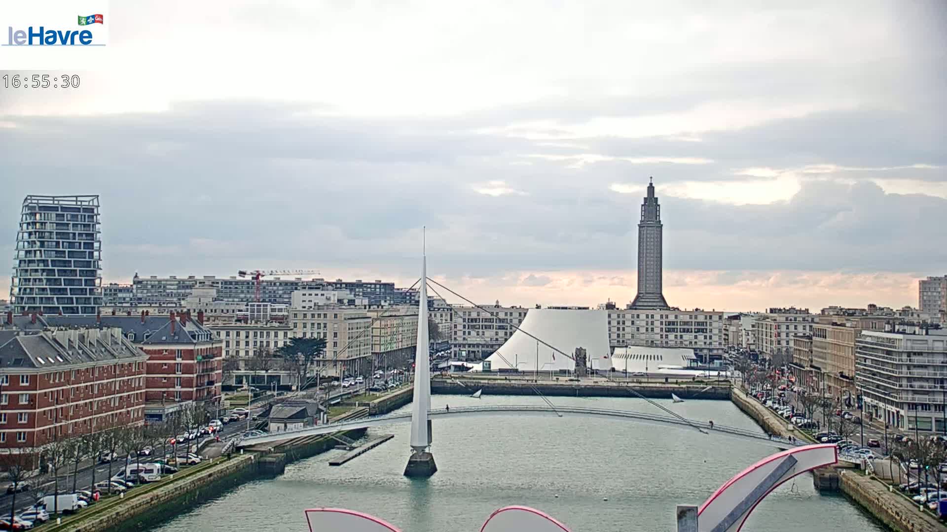 A cityscape with overcast skies shows a body of water, a modern pedestrian bridge, several buildings, and a tall tower.