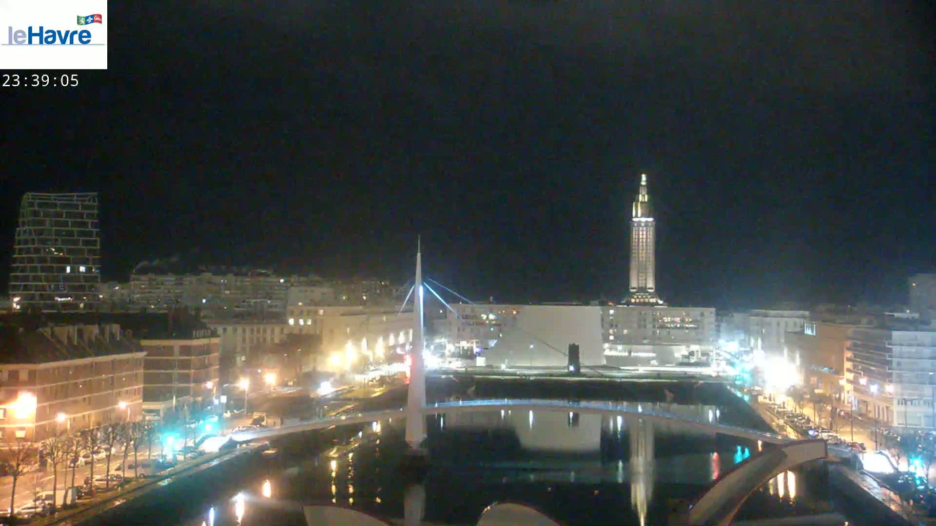 A nighttime view of a city with a bridge, a tall tower, and buildings illuminated along a waterway under a dark sky.