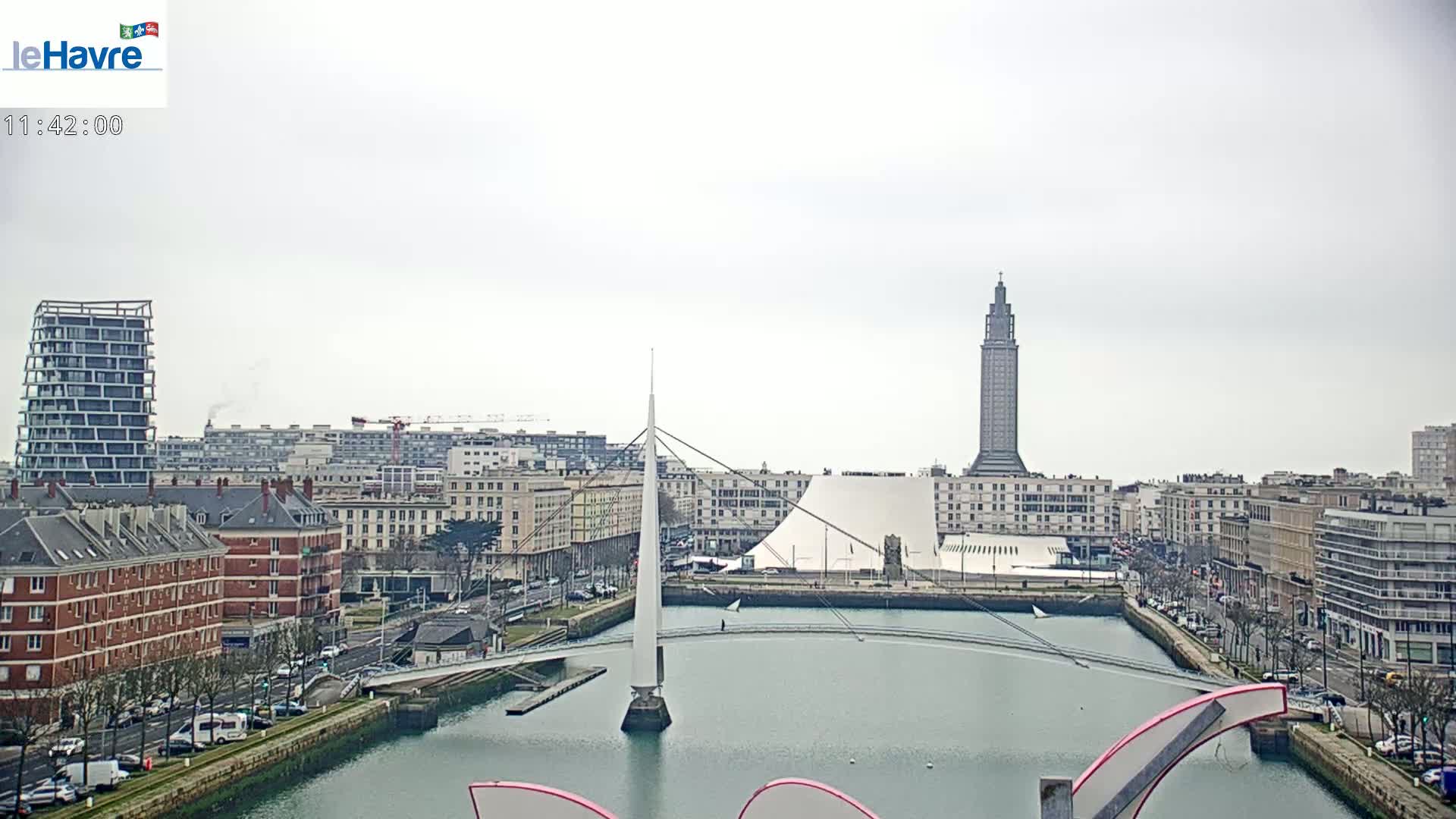 A cityscape under an overcast sky shows a waterway with a modern bridge and buildings, including a tall tower.