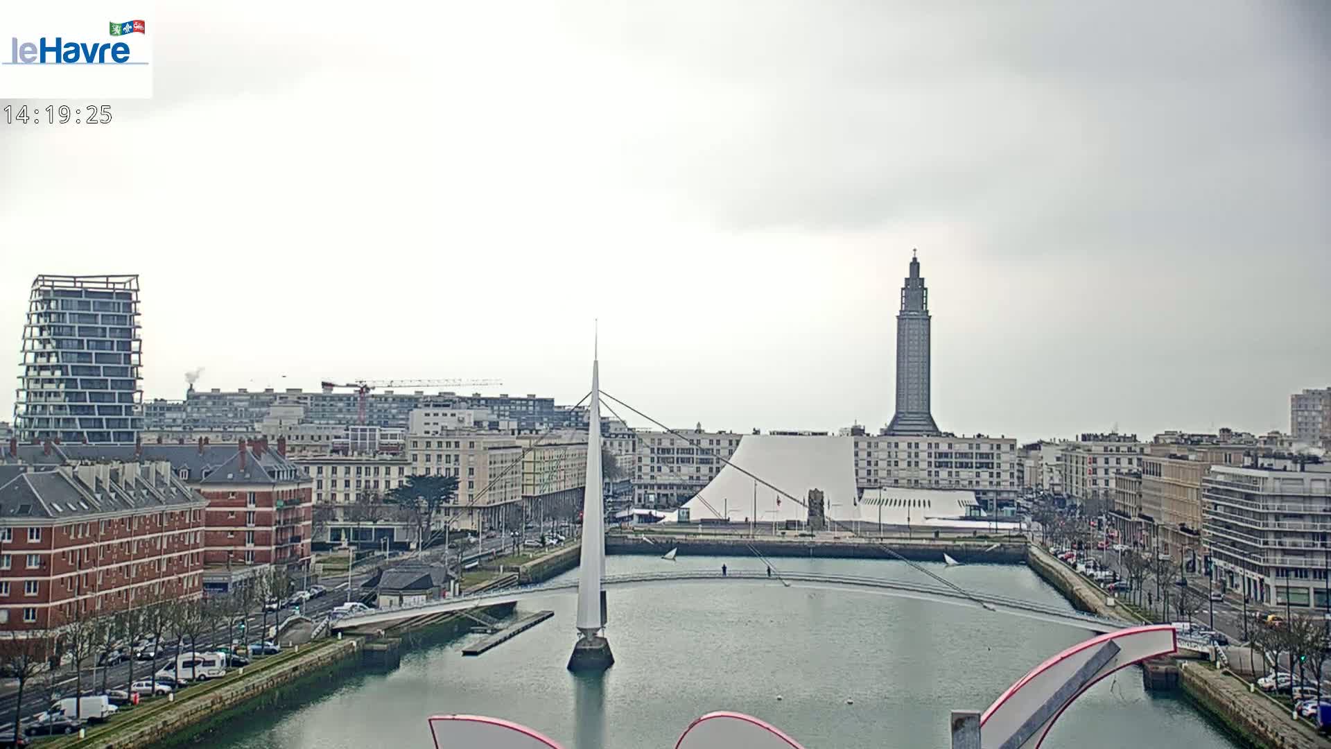 A city waterfront scene on an overcast day shows a canal with a modern bridge and several buildings, including a tall tower and a large white domed structure.