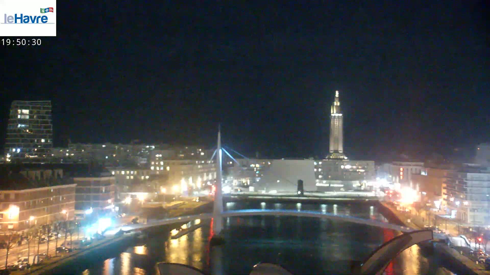 A nighttime view of a city waterfront, showing a bridge, buildings with illuminated windows, and a tall tower, under a clear, dark sky.
