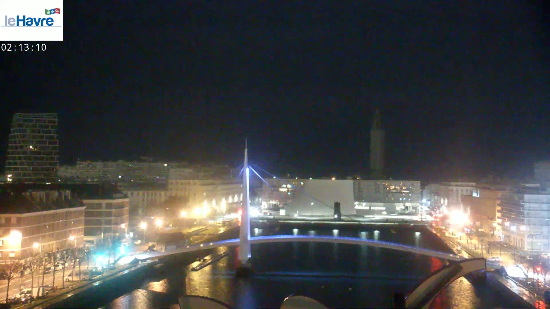 A nighttime view of a city with a lit bridge spanning a waterway, buildings lining the waterfront, and a clear, dark sky.