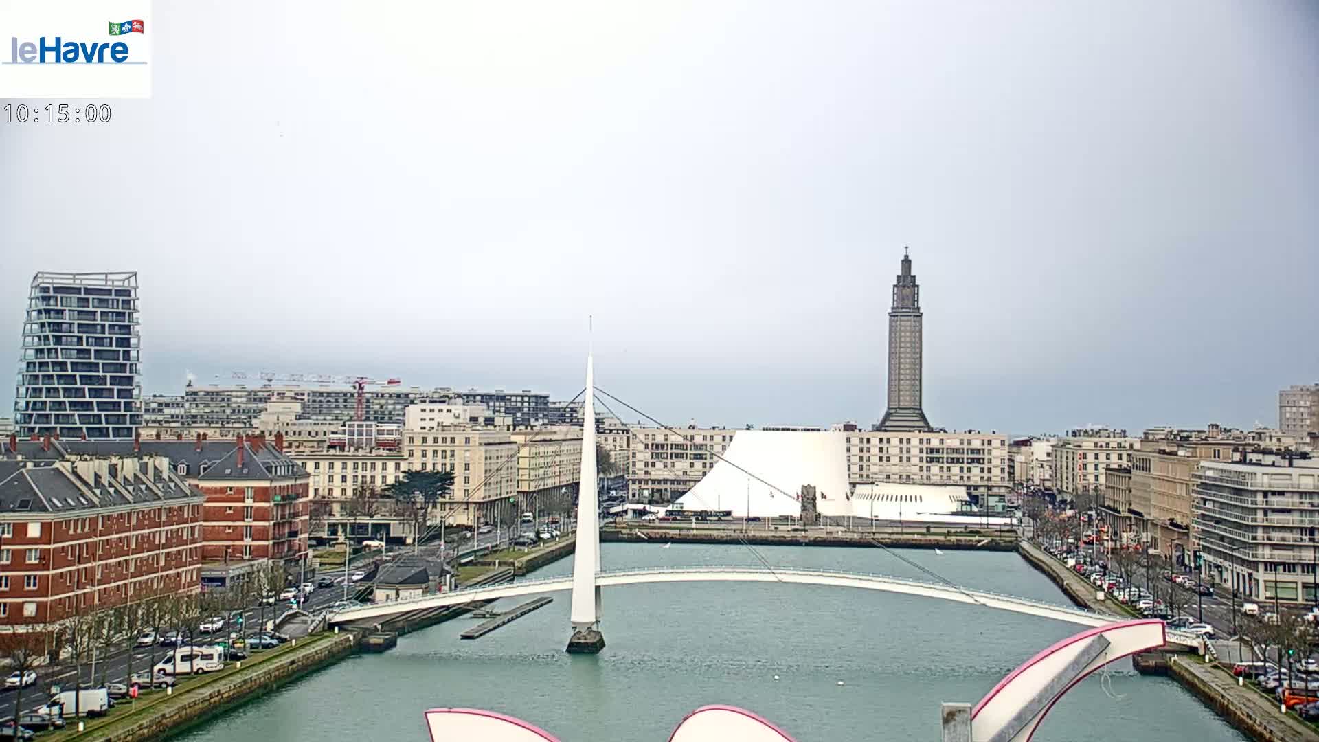 A city waterfront scene on an overcast day shows a white bridge spanning a waterway, with buildings and a tall tower in the background.