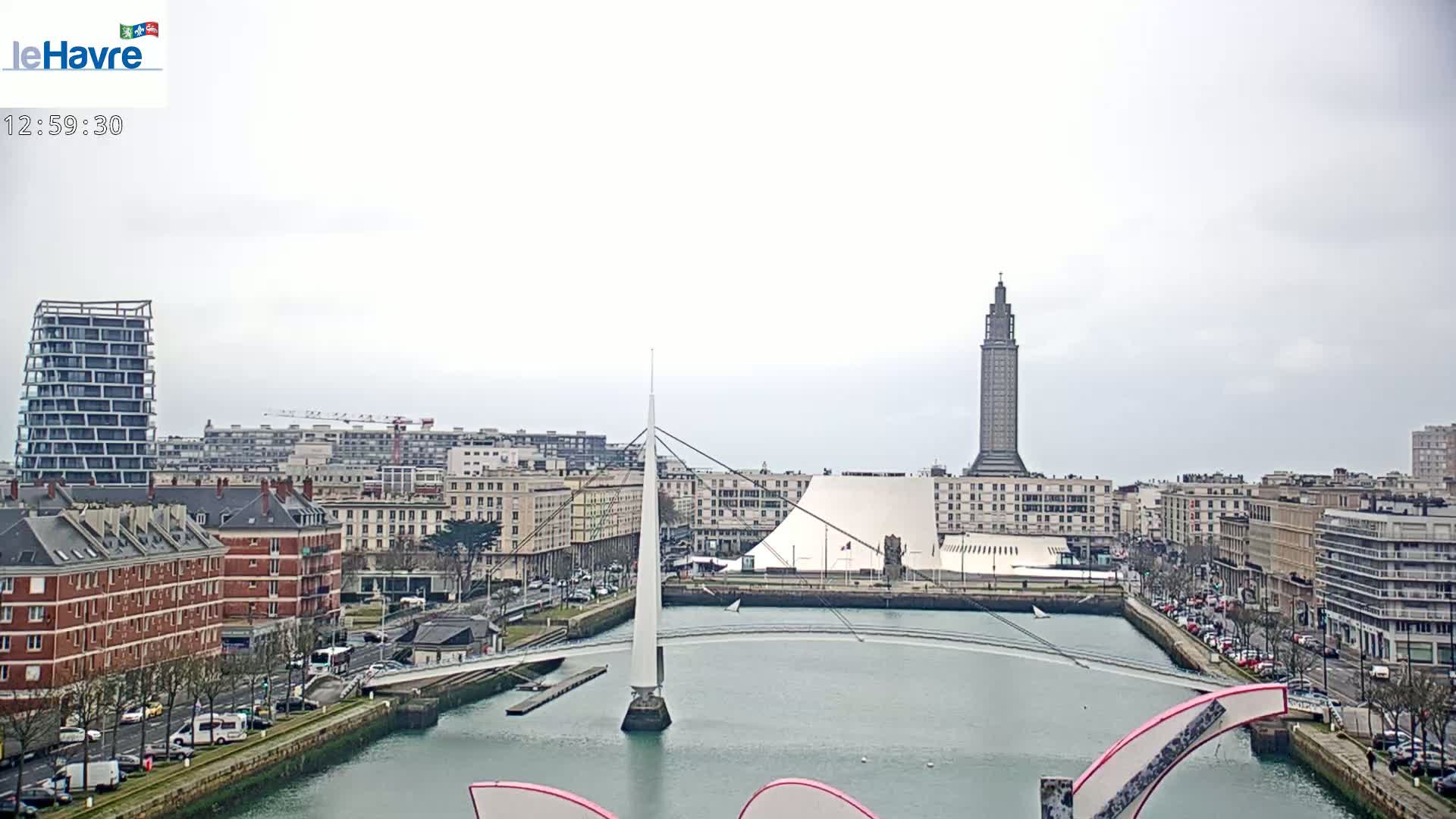 A cityscape with a tall tower, modern buildings, and a white pedestrian bridge spanning a calm body of water under an overcast sky.