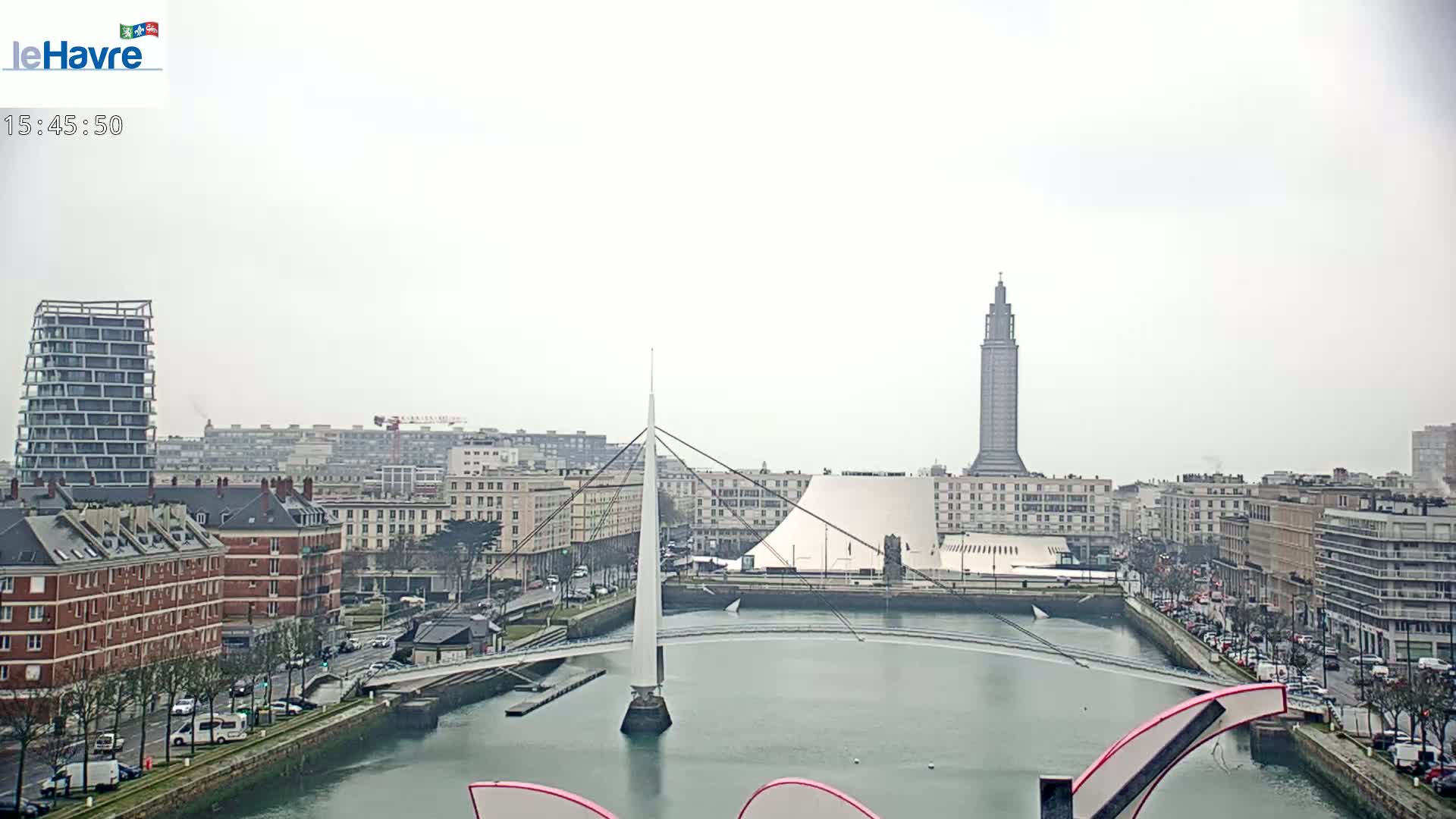 A city waterfront on an overcast day features a slender bridge, a tall tower, and modern buildings along a waterway.