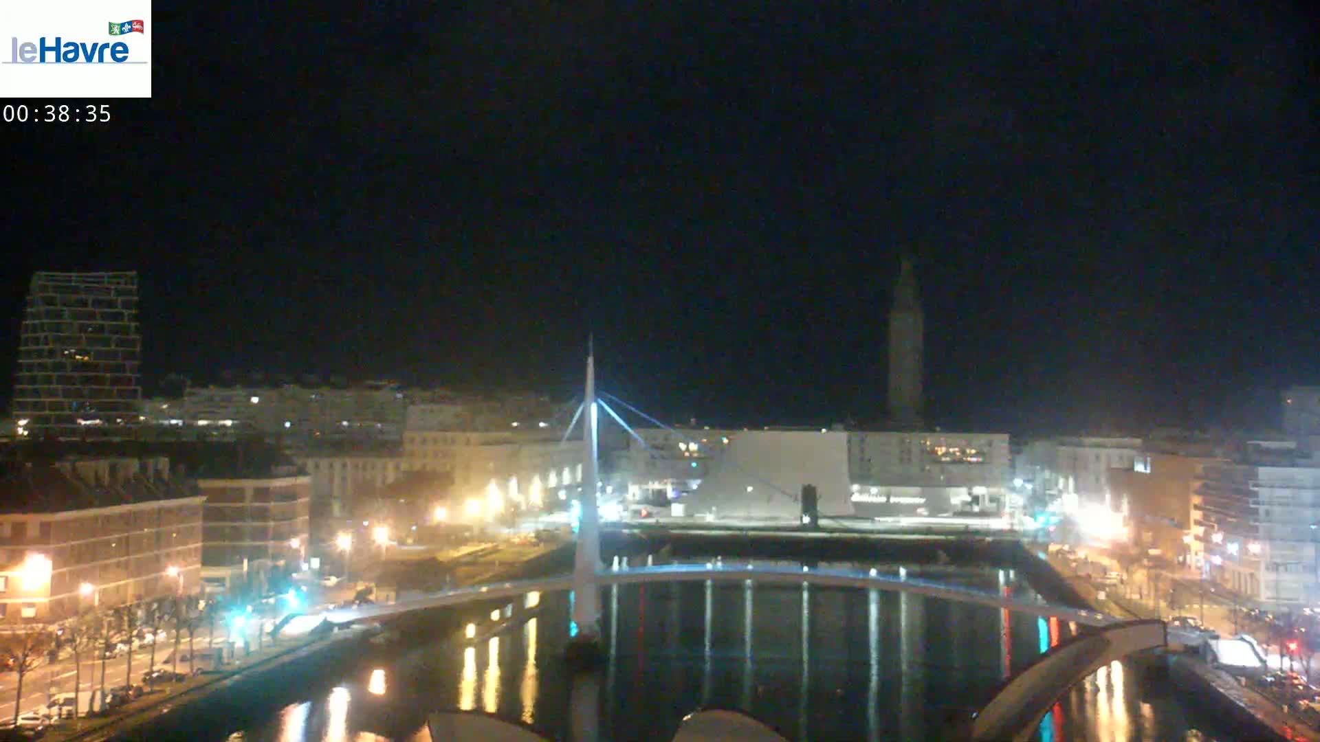 A nighttime view of a city's waterfront, showing a bridge, buildings with lights, and a tall structure in the distance.