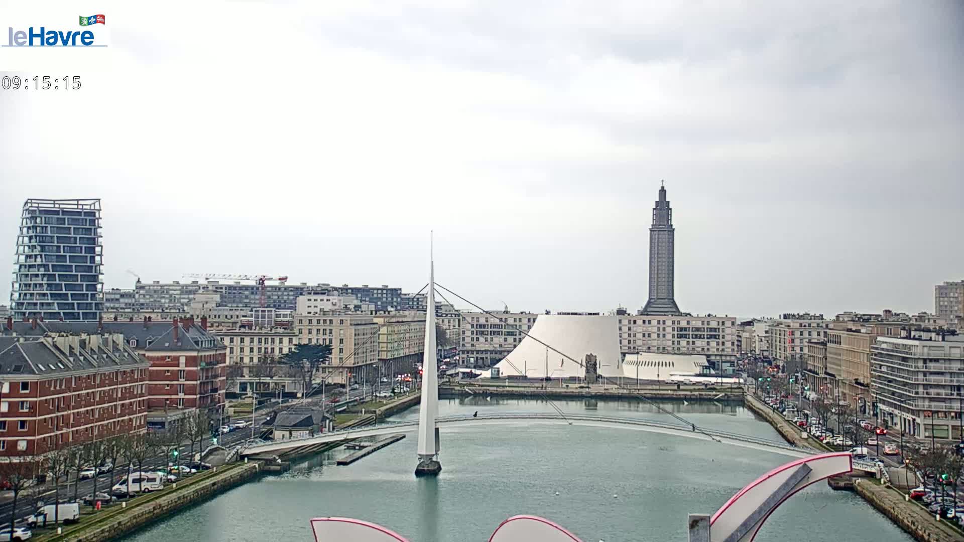 A city waterfront on an overcast day features a slender white bridge spanning a canal, with buildings and a tall tower visible in the background.