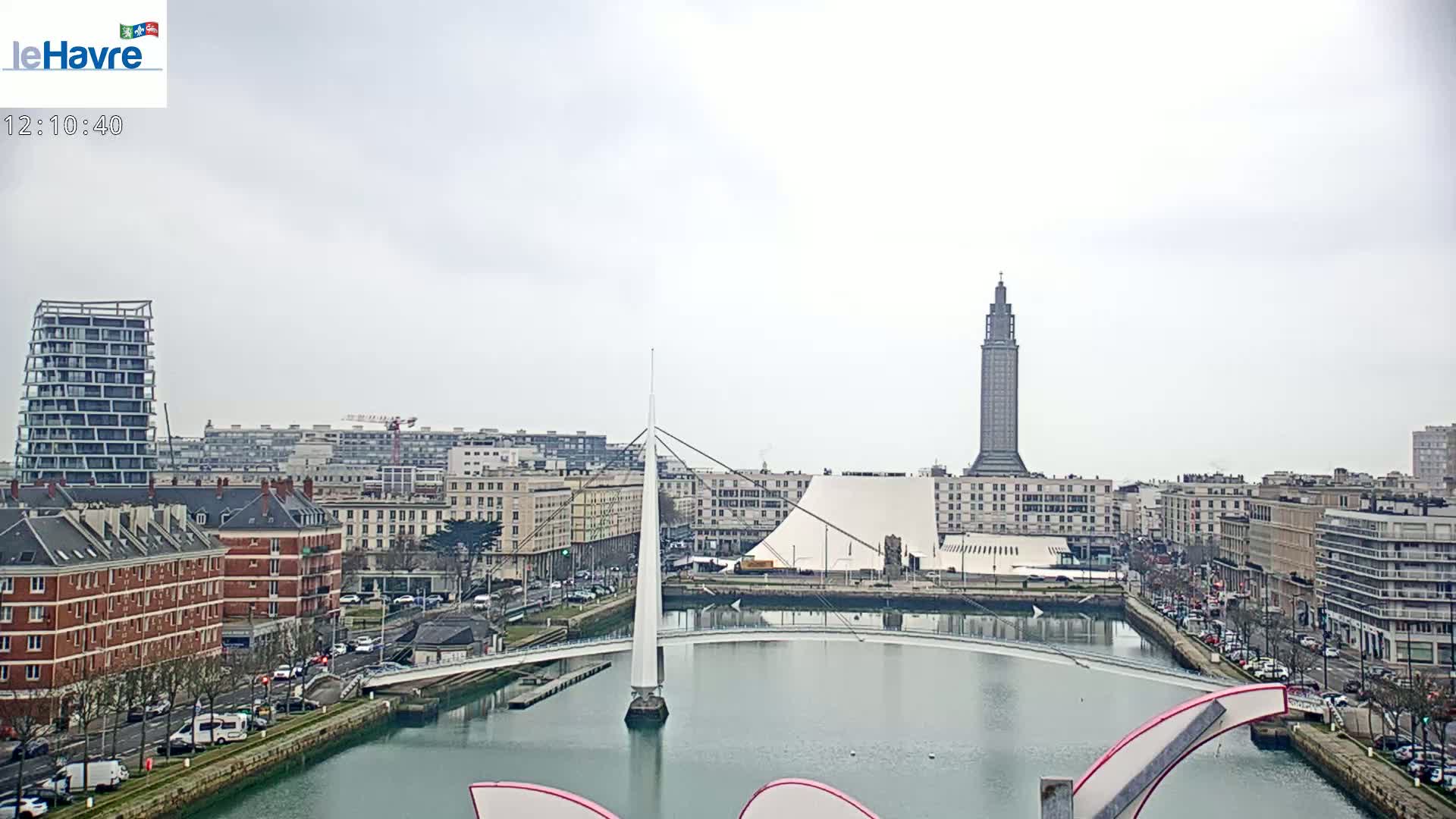 A city skyline with overcast skies shows a canal, a modern white bridge, and a tall, dark tower in the distance.