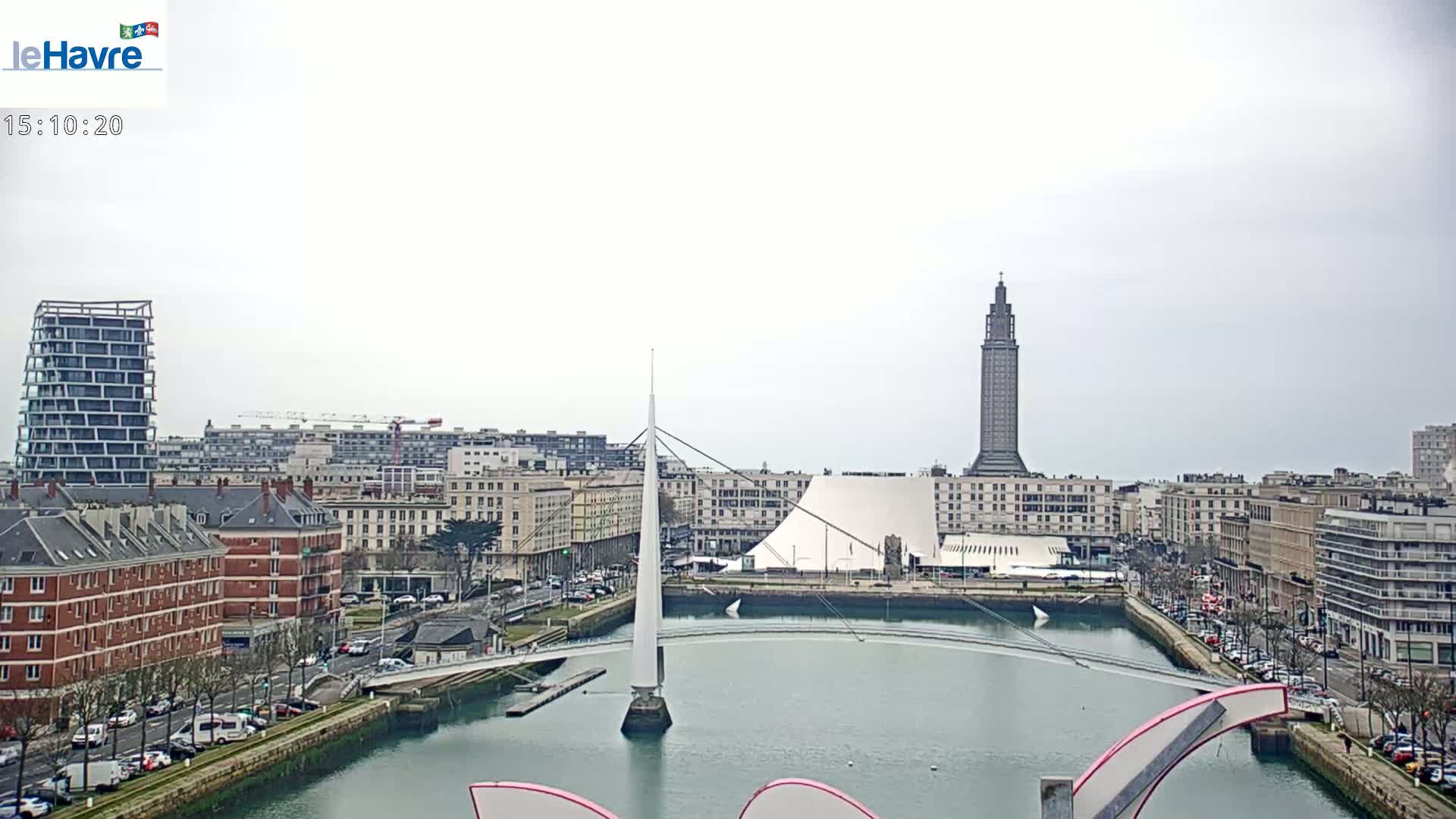 A cityscape with overcast skies shows a waterway with a modern pedestrian bridge, buildings, and a tall tower in the background.