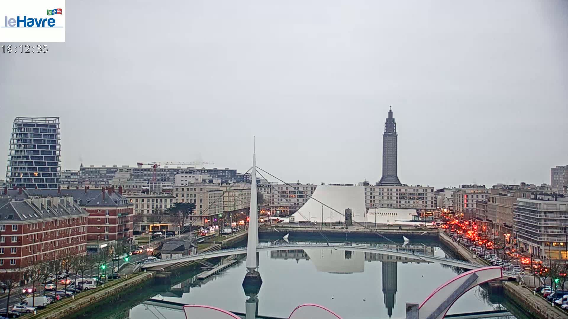 A city waterfront on an overcast day, featuring a tall tower, a modern white building, a bridge, and a canal reflecting the buildings and sky.