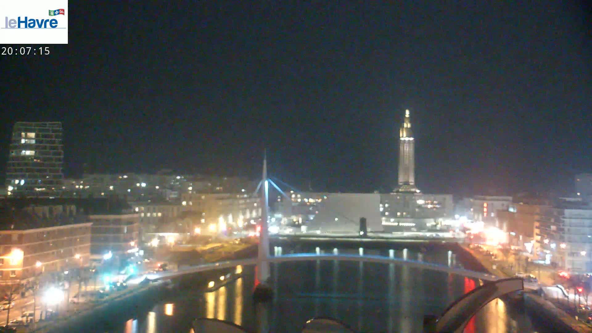 A nighttime view of a city waterfront, featuring a bridge, buildings with lights, and a tall tower in the distance under a clear, dark sky.