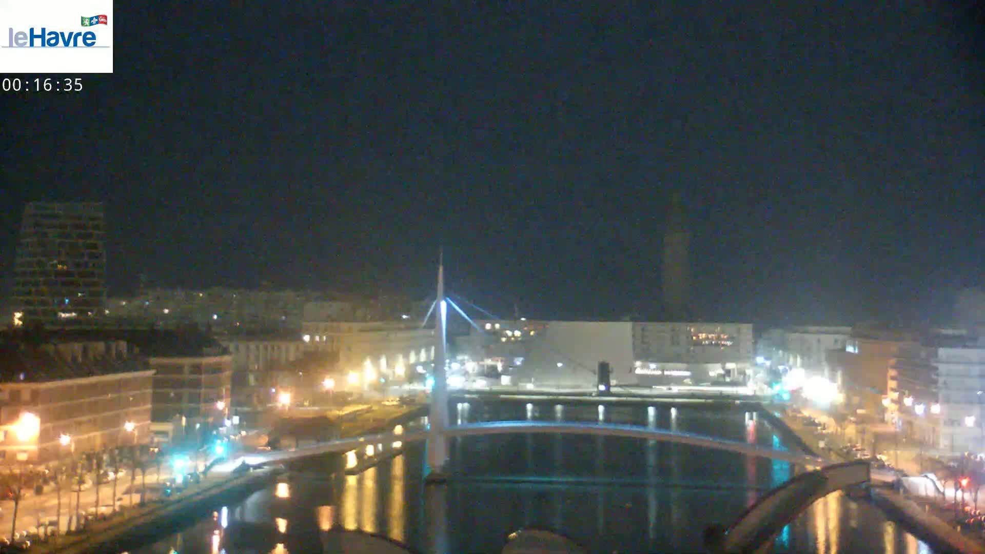 A nighttime view of a city's waterfront, featuring a modern bridge over a calm body of water, with buildings and streetlights illuminating the scene.