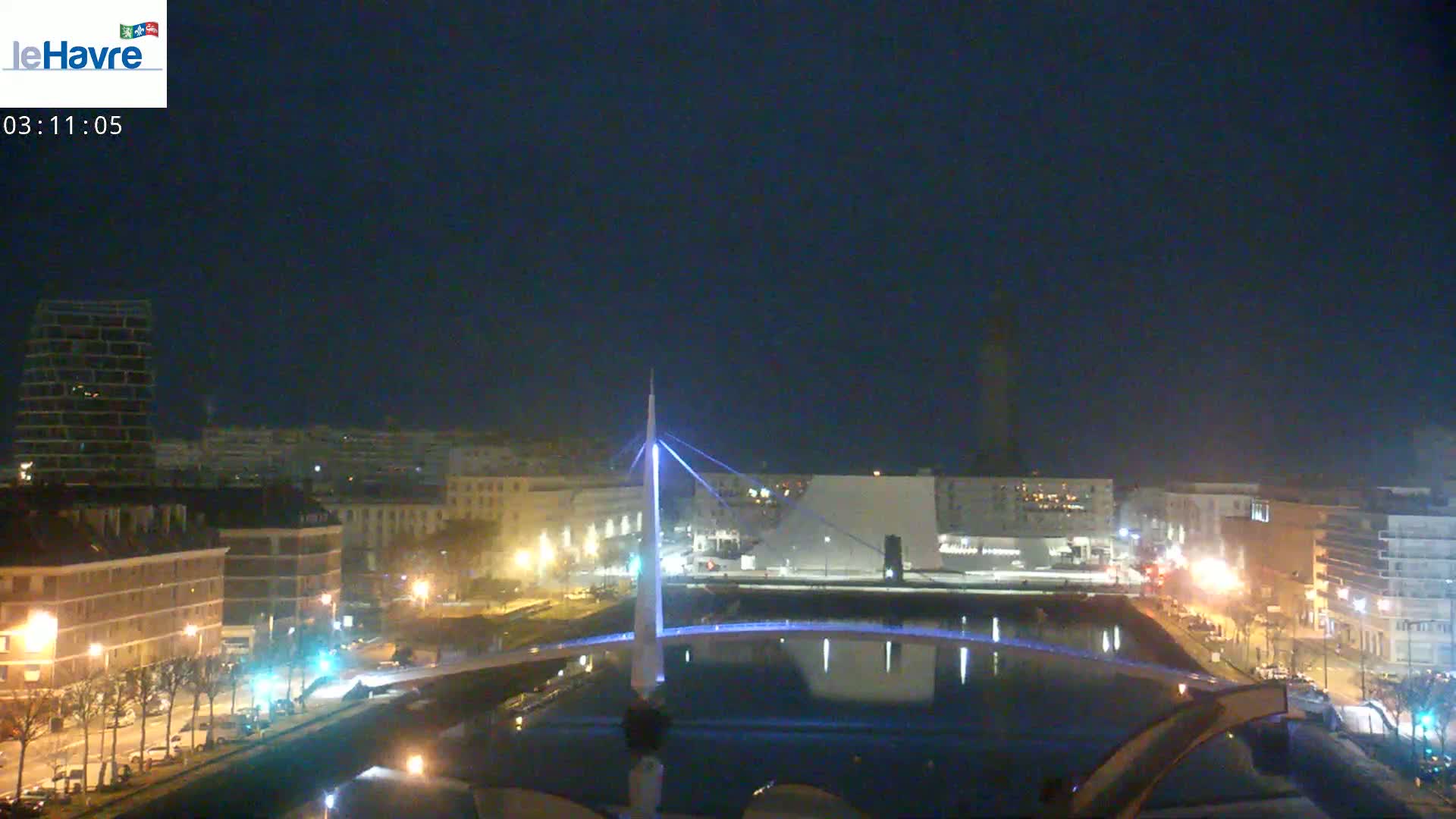 A nighttime view of a city with a lit-up bridge spanning a calm body of water, under a dark, clear sky.