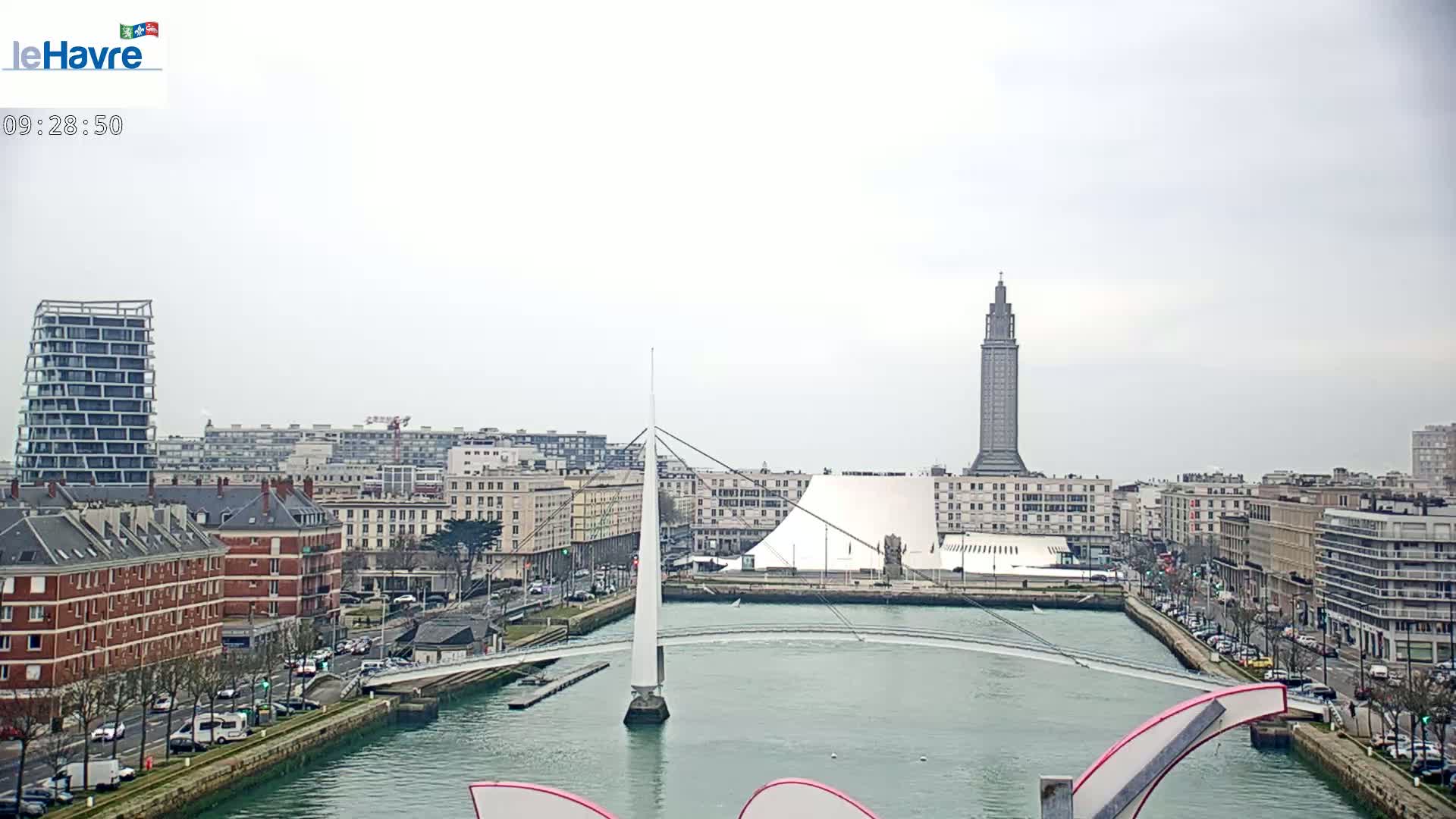 A city waterfront on an overcast day features a slender white pedestrian bridge spanning a waterway, with modern and older buildings lining the shores, including a tall tower in the background.