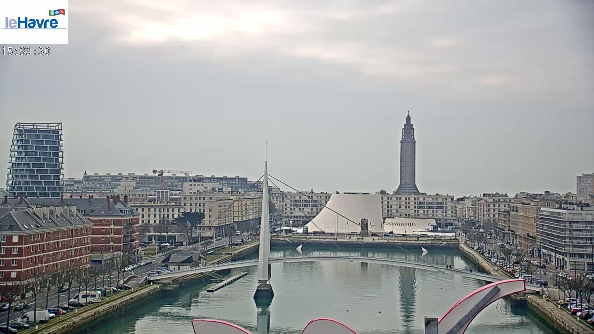 A city waterfront with overcast skies shows a canal, a bridge, several buildings, and a tall tower in the distance.