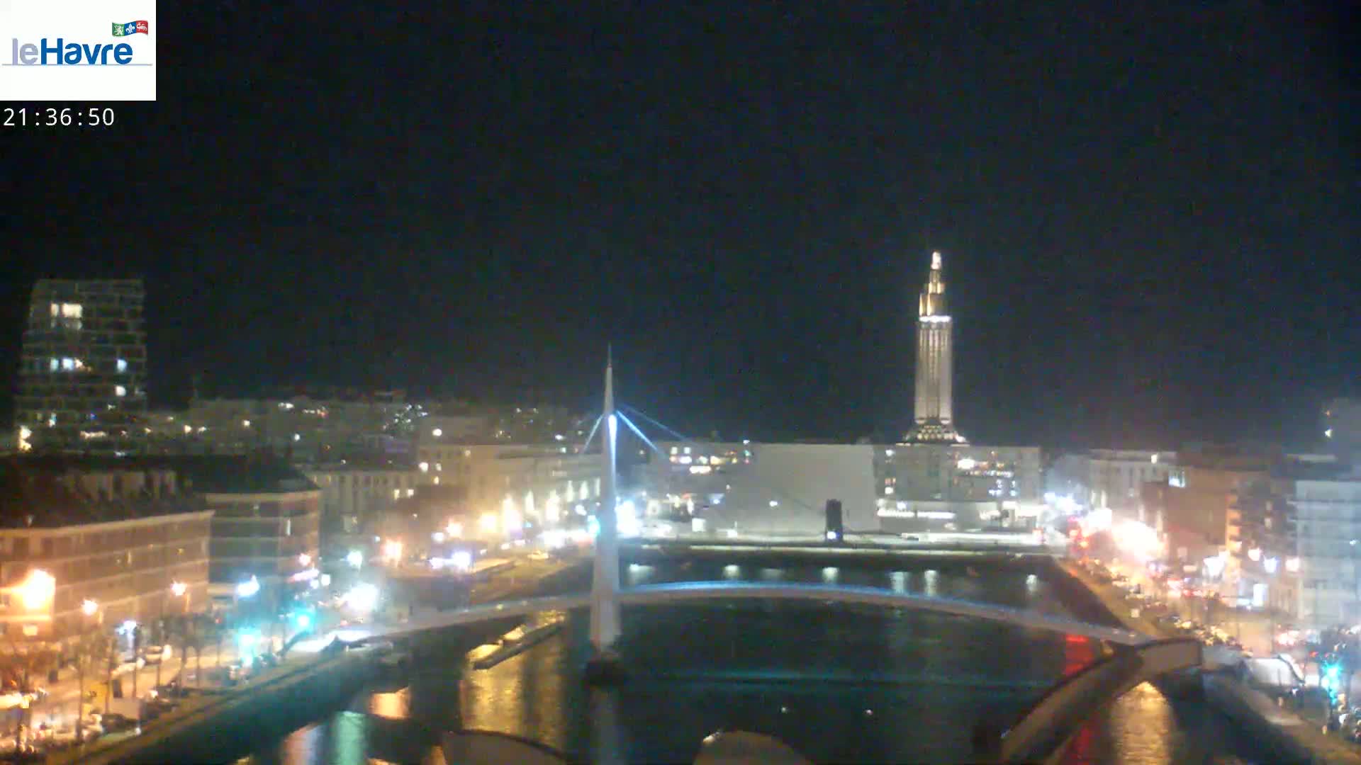 A nighttime view of a city with a bridge spanning a waterway, illuminated buildings, and a tall tower in the background under a clear, dark sky.