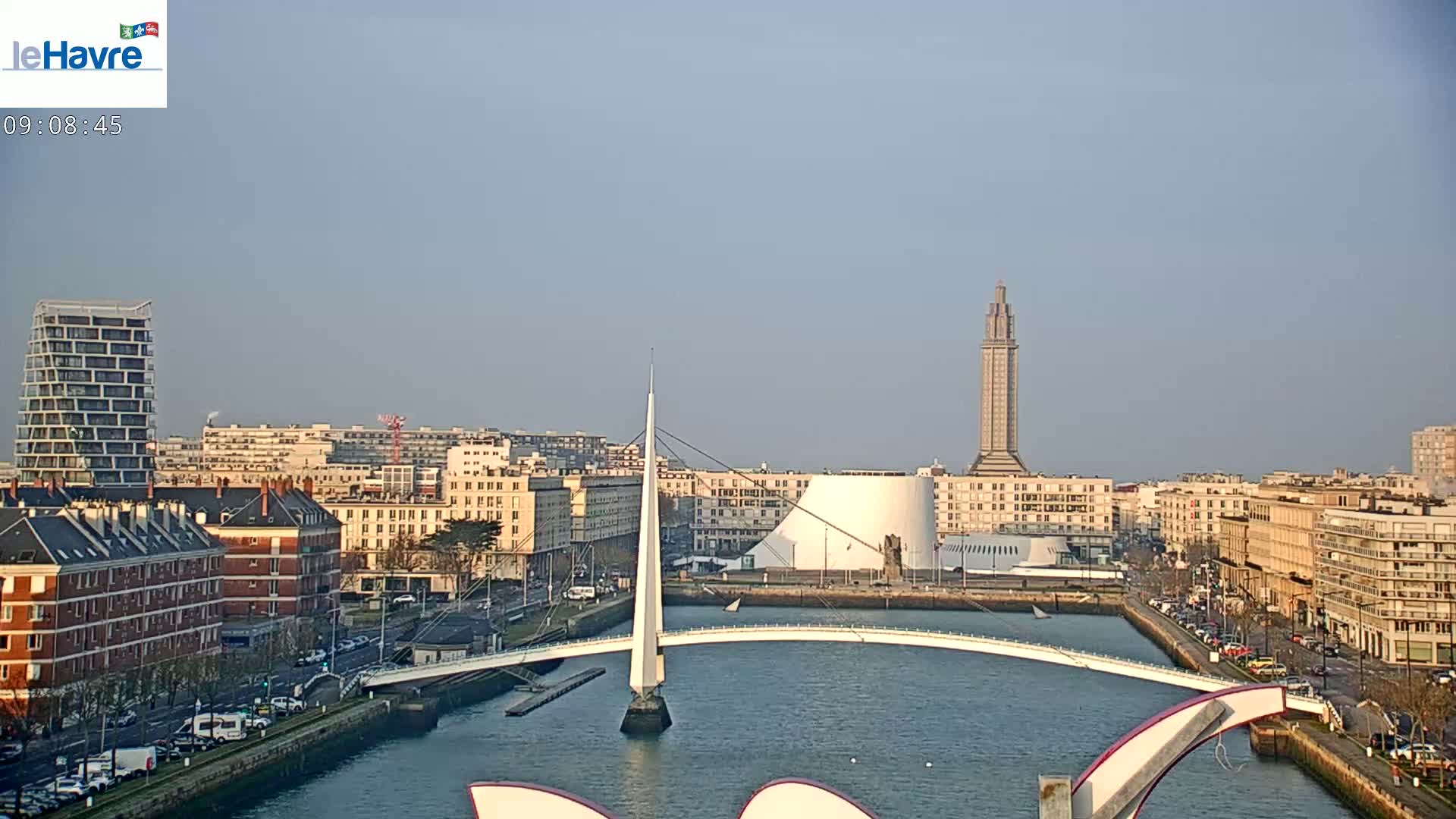 A city skyline with a modern white bridge spanning a waterway, under a clear, light blue sky.