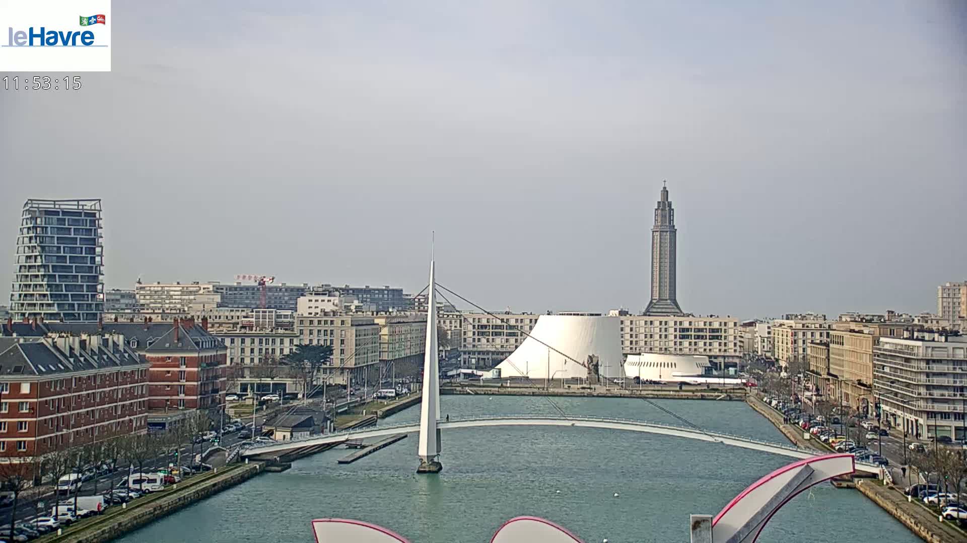 A city waterfront on a cloudy day, featuring a slender bridge, a tall tower, and modern buildings along a canal.
