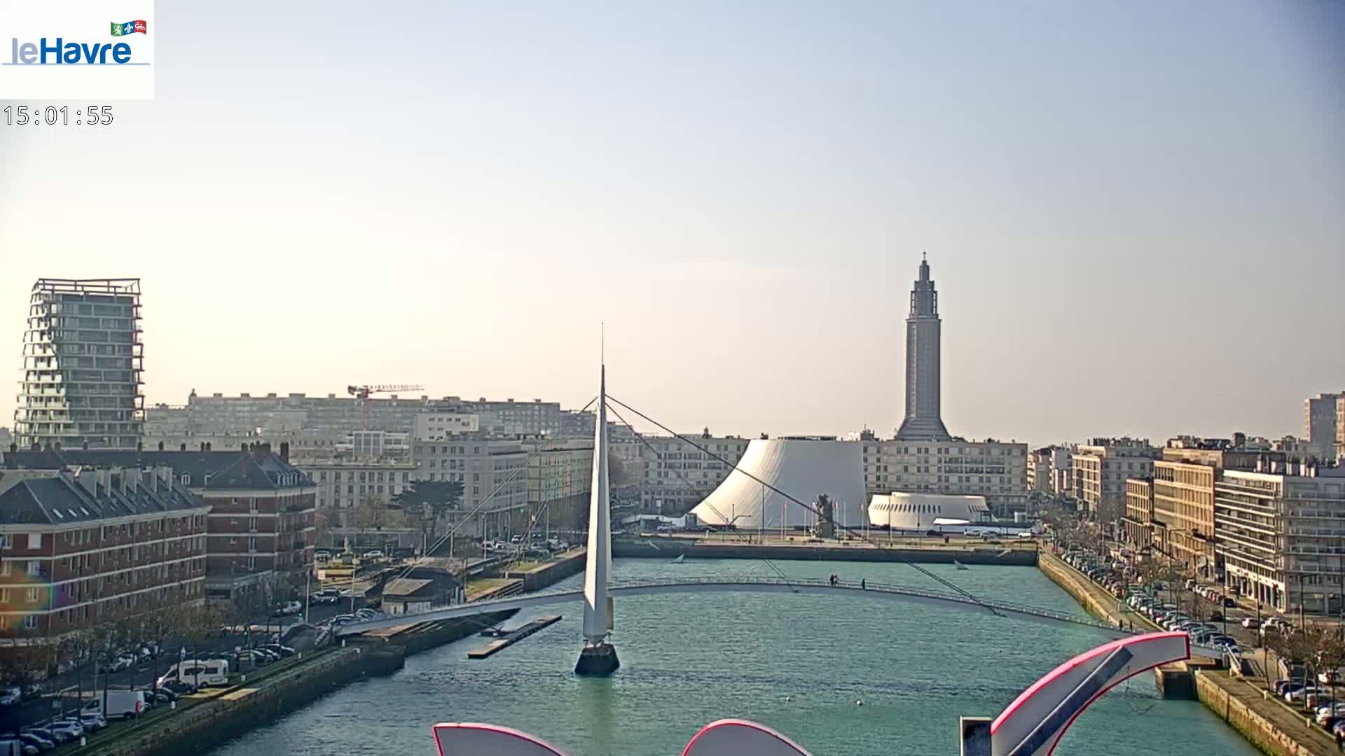 A city waterfront on a sunny day features a slender bridge, modern buildings, and a tall tower in the distance.