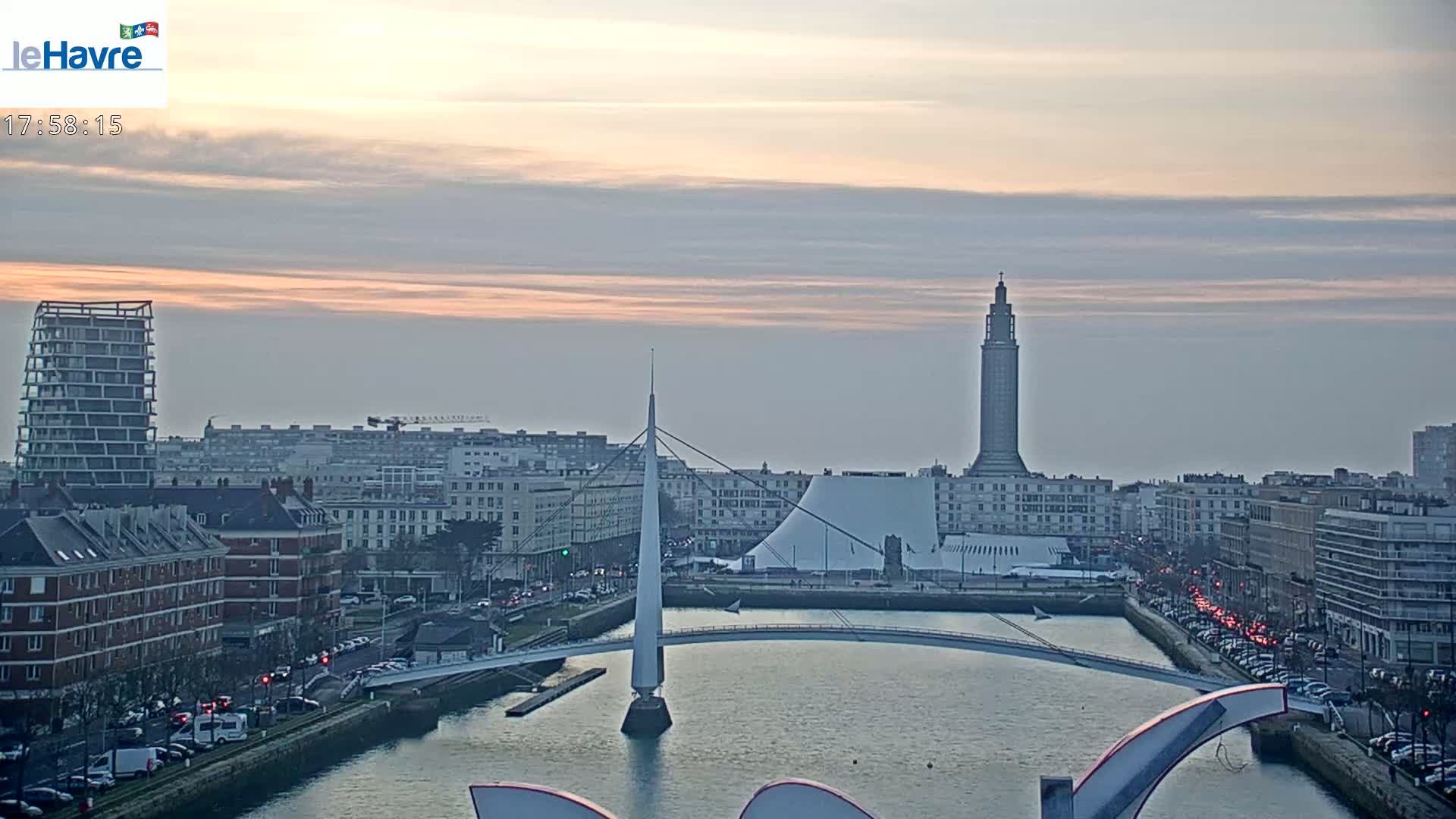 A city skyline at sunset, featuring a tall tower, modern buildings, a bridge over a waterway, and a partly cloudy sky.